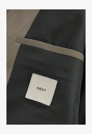 Schwarzer und beige Stoff mit einem strukturierten hexagonalen Muster, versehen mit einem kleinen Etikett mit "NEXT" in schwarz auf weißem Hintergrund und einem kontrastierenden Rand.