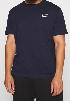 Camiseta de algodón azul marino con cuello redondo, mangas cortas y un logo "LACOSTE" blanco bordado con un emblema de cocodrilo en el pecho.