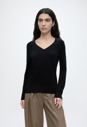 Mujer con cabello largo y oscuro, vistiendo un suéter negro de cuello en V y pantalones beige holgados, de pie sobre un fondo blanco liso.