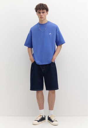 Jeune homme debout contre un mur blanc portant un t-shirt bleu oversize, un short en jean foncé, des chaussettes blanches, des baskets noires et blanches, et un collier.