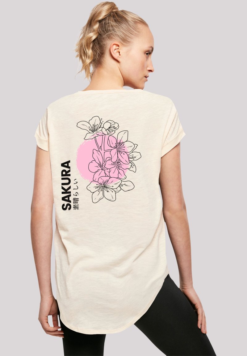 Cremfarbendes T-Shirt mit kurzen Ärmeln und floralem Design in Schwarz und Pink auf der Rückseite. Der Text lautet "SAKURA" in fetten schwarzen Buchstaben. Weicher Stoff.