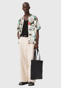 Chemise à manches courtes à motif floral sur fond blanc, associée à un débardeur noir et un pantalon large beige. Sac cabas noir, chaussures noires brillantes.