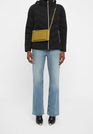 Osoba nosząca czarną kurtkę puchową, jasnoniebieskie jeansy o prostych nogawkach, czarne botki z metalowymi noskami oraz oliwkową torbę przez ramię.