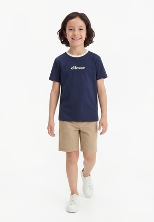 Χαμογελαστό παιδί με σκούρα σγουρά μαλλιά που περπατά προς τα εμπρός, φορώντας μπλε μπλε "ellesse" μπλουζάκι, μπεζ σορτς και άσπρα αθλητικά παπούτσια σε λευκό φόντο.