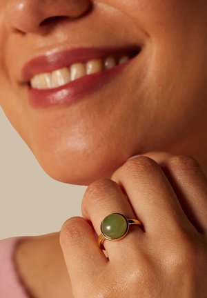 Personne souriante à la peau de ton moyen portant une bague en or avec une pierre ronde verte sur le doigt, près du menton.