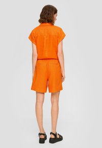 Oranžový lněný dvoudílný outfit, který zahrnuje košili s krátkým rukávem a odpovídající šortky s elastickým pasem a zadními kapsami. Černé sandály.