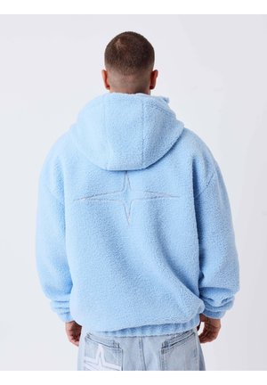 Mann trägt hellblaue Fleecejacke mit Kapuze und sternförmiger Naht auf dem Rücken, Blick weg vor weißem Hintergrund.