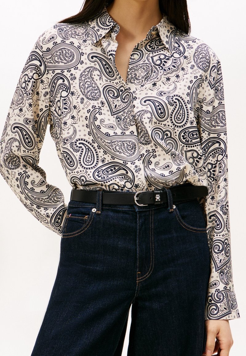 Chemise blanche à manches longues avec motifs cachemire noirs rentrée dans un jean bleu foncé, portée avec une ceinture noire avec une boucle argentée.