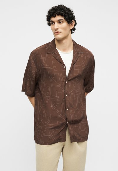 Νέος άνδρας με σγουρά μαλλιά που φορά καφέ πουκάμισο με κοντά μανίκια και σχέδια πάνω από λευκό T-shirt και μπεζ παντελόνι, κοιτάζει αριστερά.