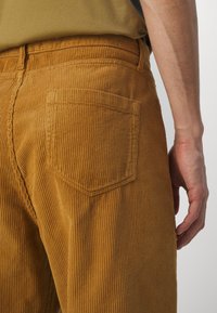 Pantalones de pana marrón con ribete vertical, que cuentan con un bolsillo trasero y un ajuste ceñido en la cintura, complementados por una superficie suave y texturada.