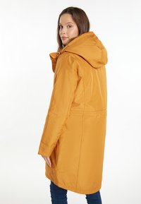 Cappotto lungo giallo con cappuccio, texture liscia e vita strizzata. Presenta una chiusura con zip e tasche laterali, su uno sfondo bianco.