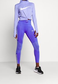 Fialová Nike zipová mikina s uzlem vpředu, doplněná o odpovídající legíny s vysokým pasem a květinovým akcentem na boku. Černé tenisky.