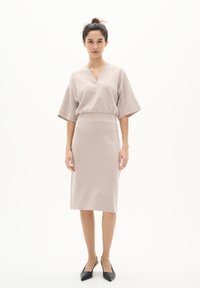 Beiges, tailliertes Kleid mit V-Ausschnitt und kurzen Ärmeln. Glatte Textur, knielang, kombiniert mit schwarzen, spitzen Ballerinas. Einfaches Design, ohne Muster.