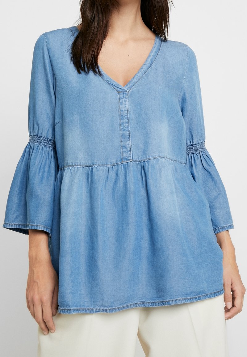 Light blue denim top with a V-neck, button detail, and flared sleeves. Features smocking at the cuffs and a gathered waist for a relaxed fit.