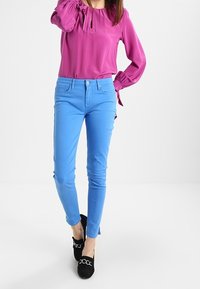 Blusa rosa de manga larga con escote en forma de ojo de cerradura, combinada con pantalones ajustados de color azul claro y zapatos negros con un acento de cadena.