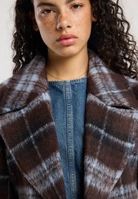 Brown and plaid wool coat with a notched collar and textured surface, layered over a blue denim top with central zipper.