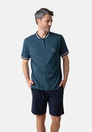 Homme portant un polo rayé bleu sarcelle à manches courtes avec un short foncé, debout les mains dans les poches, regardant vers sa droite.
