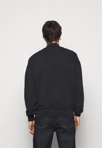 Black sweatshirt with a ribbed collar and cuffs, featuring a relaxed fit and smooth texture. Worn over dark denim pants.