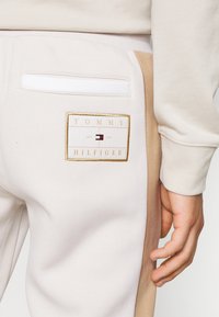 Lys beige sweatpants med et kontrasterende tan sidepanel. Har en hvid lomme og et guldbroderet logo-patch på bagsiden.