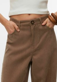 Pantalon en velours côtelé marron taille haute, fermeture à boutons sur le devant et poches latérales, avec une texture côtelée et une coupe ajustée.