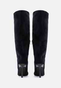 Langsvarte høye støvler i semsket skinn med tettsittende design, glidelåsdetaljer på sidene og slanke høye hæler. Glatt tekstur og slank silhuett.