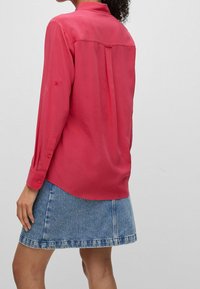 Fuchsia Langarmhemd mit Kragen, knöpfen an den Bündchen und einer Rückenfalte, kombiniert mit einem hellblauen Jeansrock mit ausgefranstem Saum.