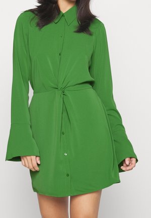 Frau trägt ein langärmeliges grünes Kleid mit Knöpfen und einer Knotendetail am Taillenbund vor einem einfarbigen Hintergrund.