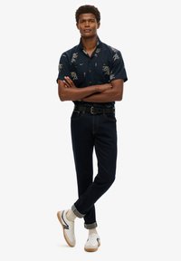 Camisa de manga corta de color navy con estampado floral, jeans oscuros, cinturón negro, zapatillas beige y calcetines claros, exhibiendo un ajuste relajado y un estilo casual.