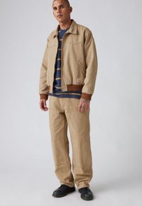 Μπεζ μπουφάν με καφέ λεπτομέρειες, συνδυασμένο με παντελόνι σε ταιριαστή μπεζ απόχρωση. Οι στρώσεις αποκαλύπτουν ένα μπλε σκούρο ριγέ πουκάμισο από κάτω. Μαύρα παπούτσια ολοκληρώνουν την εμφάνιση.