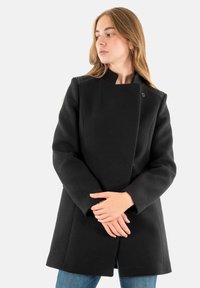 Zwarte wollen jas met een hoge kraag, voorzien van decoratieve stiksels op de schouders, een enkele knoopsluiting en een getailleerde pasvorm.