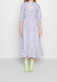 Žena v levandulových květinových midi šatech s volánkovým límečkem, tříčtvrtečními rukávy a světle zelenými kotníkovými botami stojící na pozadí bílé stěny.