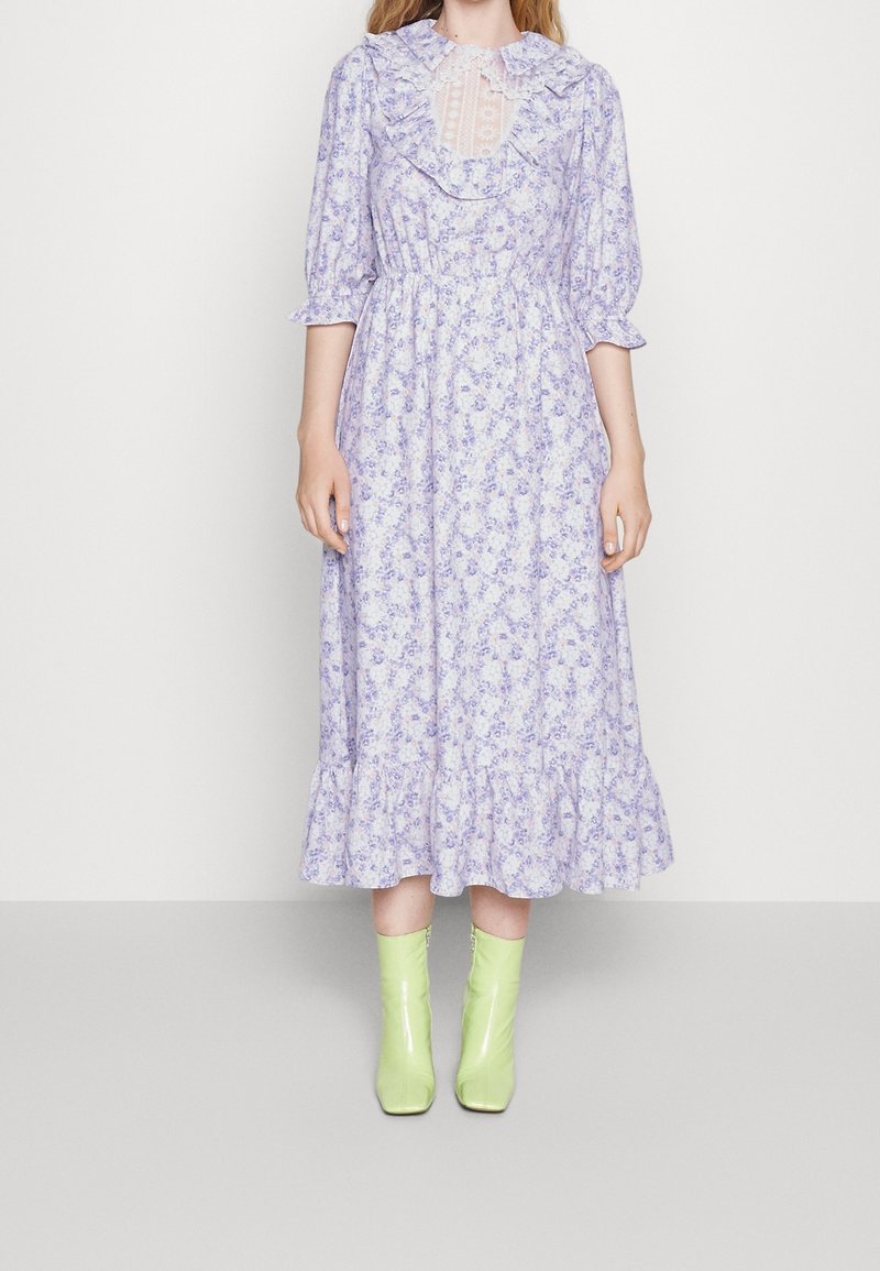 Žena v levandulových květinových midi šatech s volánkovým límečkem, tříčtvrtečními rukávy a světle zelenými kotníkovými botami stojící na pozadí bílé stěny.
