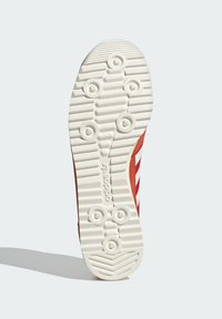 Semelle de chaussure de sport rouge avec un motif de traction en caoutchouc blanc, présentant des rainures circulaires et linéaires, et le logo "adidas" embossé près du talon.