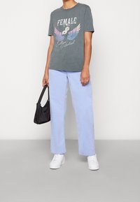 Harmaa graafinen t-paita, jossa on siivet ja teksti, vaaleansiniset suorat farkut, valkoiset tennarit ja pieni musta käsilaukku. Raskaat pinnat.