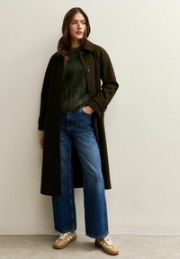 Vrouw staand tegen een beige muur, gekleed in een donkergroene jas, een groene zachte trui, blauwe wijd-uitlopende jeans en beige sneakers met witte veters.