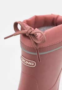 Botte en caoutchouc rose avec une surface texturée, des accents gris et un col en tissu noué. Logo visible sur le côté. Présente une forme de haut arrondi.