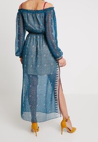 Niebieska przezroczysta maxi sukienka z dekoltem off-the-shoulder, długimi rękawami, kwiatowym wzorem, podkreśloną talią i bocznym rozcięciem; zestawiona z żółtymi szpilkami.