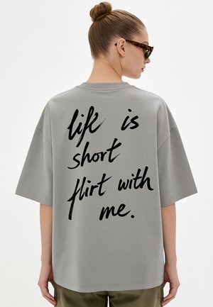 Donna con capelli raccolti in uno chignon, che indossa una T-shirt grigia oversize con testo nero "life is short flirt with me" sulla schiena e occhiali da sole scuri.