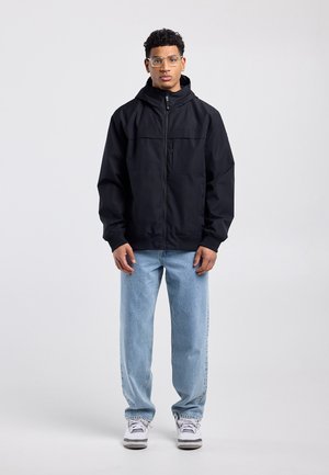 Junger Mann mit klaren Brillengläsern, schwarzer Jacke mit Reißverschluss und Kapuze, hellblauen Jeans und weißen Turnschuhen, stehend vor schlichtem grauem Hintergrund.