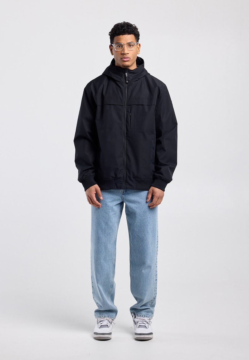 Junger Mann mit klaren Brillengläsern, schwarzer Jacke mit Reißverschluss und Kapuze, hellblauen Jeans und weißen Turnschuhen, stehend vor schlichtem grauem Hintergrund.