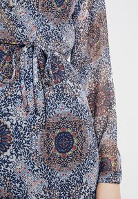 Close-up van een doorschijnende, gemusterde jurk met een multikleurige bloemen- en geometrische print, gestrikt in de taille met een bijpassende stoffen riem.