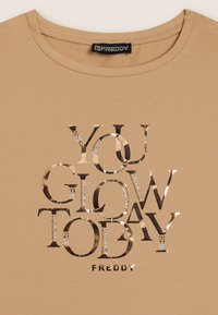 Camiseta de algodón beige con letras metálicas en color oro rosa que dicen "HOY BRILLAS" con toques de strass; cuello redondo.