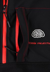 Vêtement noir avec fermeture éclair et coutures rouges, présentant une poche ornée d'un design brodé de lignes entremêlées blanches et du texte rouge "Rivūn PROJECTS".