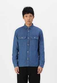 Denim button-up shirt in medium blue, features two chest pockets, gold buttons, and long sleeves with cuff buttons. Smooth texture.