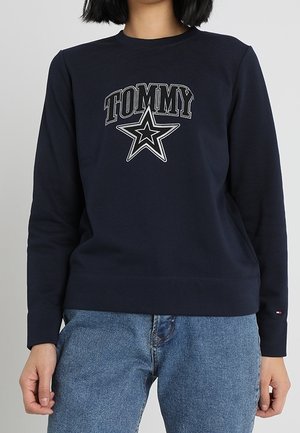 Persoon draagt een navyblauwe trui met "TOMMY" en een sterrenlogo, gecombineerd met een blauwe spijkerbroek.