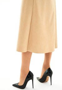 Black patent leather high heels with pointed toes and stiletto heels, worn with a light beige textured midi skirt.