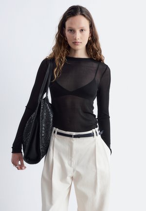 Femme aux longs cheveux bouclés portant un haut noir transparent à manches longues, un pantalon blanc taille haute, portant un grand sac cabas noir tressé sur l'épaule.