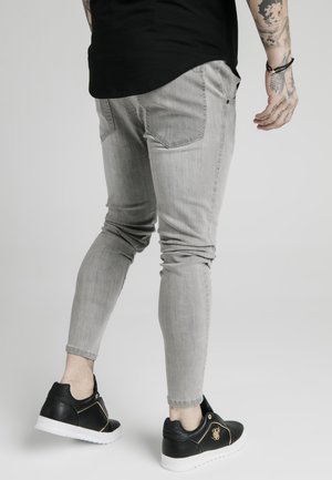 Jeans de corte slim gris con un acabado desgastado, combinados con zapatillas de cuero negras con detalles dorados y un cuello texturizado.