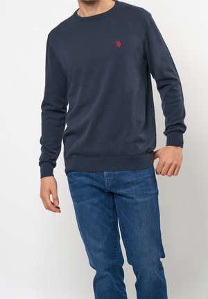Mann in dunkelblauem Langarmpullover mit rotem Logo auf der Brust und blauen Jeans, der vor einem schlichten hellen Hintergrund steht.