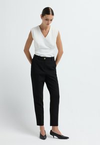 Witte mouwloze top met een V-hals, gecombineerd met zwarte getailleerde broek met een gouden knoop, en zwarte puntige hakken.